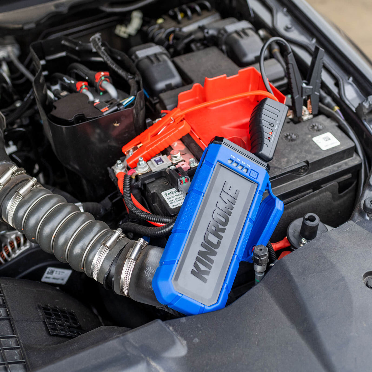 This is an image of a Kincrome automotive battery jump starter inside a car engine bay, featuring red jumper cables, from Kennedys Welding Supplies in Derwent Park TAS, tool shop and welding specialists.