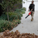 This is an image of a man using a red leaf blower on a concrete driveway with garden plants nearby, showcasing Kennedys Welding Supplies in Derwent Park TAS, offering tool shop, power tool repairs, welding specialists