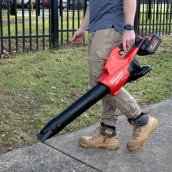 This is an image of a person using a Milwaukee cordless blower on a sidewalk with grass and a fence at Kennedys Welding Supplies in Derwent Park TAS, offering tool shop and power tool repairs